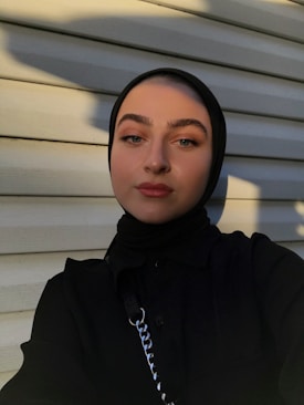 A person wearing a dark hijab and black clothing is standing in front of a light-colored wooden wall. The shadows cast by the sunlight create interesting patterns on the wall and the person's face.