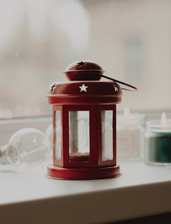 A red metal lantern with star-shaped cutouts is placed on a white surface. In the background, there are two lit candles, one to the right of the lantern and another behind it. An overturned glass bulb lies beside the lantern on the left. The background is softly blurred, suggesting a calm indoor setting.