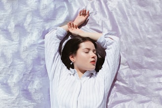 A person with closed eyes lies peacefully on a shimmering, light lavender surface. They are wearing a white striped shirt, and their arms are relaxed above their head, suggesting a state of rest or meditation.
