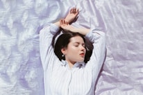 A person with closed eyes lies peacefully on a shimmering, light lavender surface. They are wearing a white striped shirt, and their arms are relaxed above their head, suggesting a state of rest or meditation.