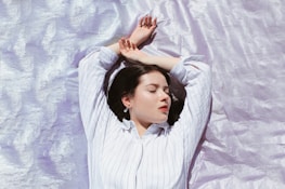 A person with closed eyes lies peacefully on a shimmering, light lavender surface. They are wearing a white striped shirt, and their arms are relaxed above their head, suggesting a state of rest or meditation.
