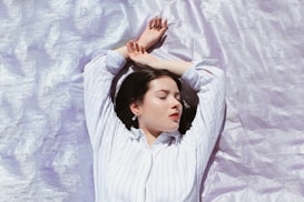 A person with closed eyes lies peacefully on a shimmering, light lavender surface. They are wearing a white striped shirt, and their arms are relaxed above their head, suggesting a state of rest or meditation.