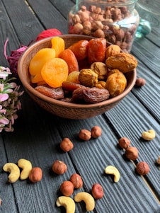 A wooden bowl filled with an assortment of dried fruits, including apricots and dates, is placed on a dark wooden surface. Scattered around the bowl are various nuts, such as hazelnuts and cashews. In the background, a glass jar is filled with more nuts, partially visible. A small bunch of pink flowers is positioned to the left of the bowl, adding a touch of color.