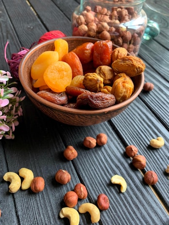 A wooden bowl filled with an assortment of dried fruits, including apricots and dates, is placed on a dark wooden surface. Scattered around the bowl are various nuts, such as hazelnuts and cashews. In the background, a glass jar is filled with more nuts, partially visible. A small bunch of pink flowers is positioned to the left of the bowl, adding a touch of color.