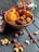 A wooden bowl filled with an assortment of dried fruits, including apricots and dates, is placed on a dark wooden surface. Scattered around the bowl are various nuts, such as hazelnuts and cashews. In the background, a glass jar is filled with more nuts, partially visible. A small bunch of pink flowers is positioned to the left of the bowl, adding a touch of color.