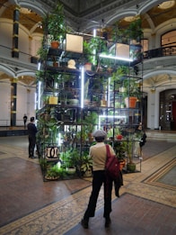 A tall metal structure filled with various potted plants is centrally placed in an ornate room with high ceilings and archways. The structure is illuminated by vertical LED lights, creating a visually striking installation. People are walking and observing the installation.