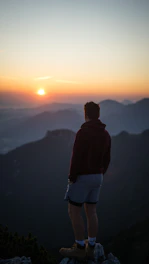 A traveler standing on a mountain peak watching the sunrise over distant landscapes.