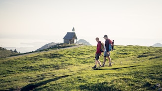 Two hikers walk along a grassy hillside towards a small, stone chapel with a pointed roof. The landscape stretches out with views of distant hills and a body of water under a bright, clear sky.
