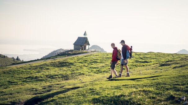 Two hikers walk along a grassy hillside towards a small, stone chapel with a pointed roof. The landscape stretches out with views of distant hills and a body of water under a bright, clear sky.
