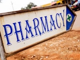 A metal sign with blue lettering indicating the direction to a pharmacy. The background is slightly blurred, showing pavement and some indistinct buildings and vehicles.