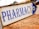 A metal sign with blue lettering indicating the direction to a pharmacy. The background is slightly blurred, showing pavement and some indistinct buildings and vehicles.