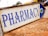 A metal sign with blue lettering indicating the direction to a pharmacy. The background is slightly blurred, showing pavement and some indistinct buildings and vehicles.