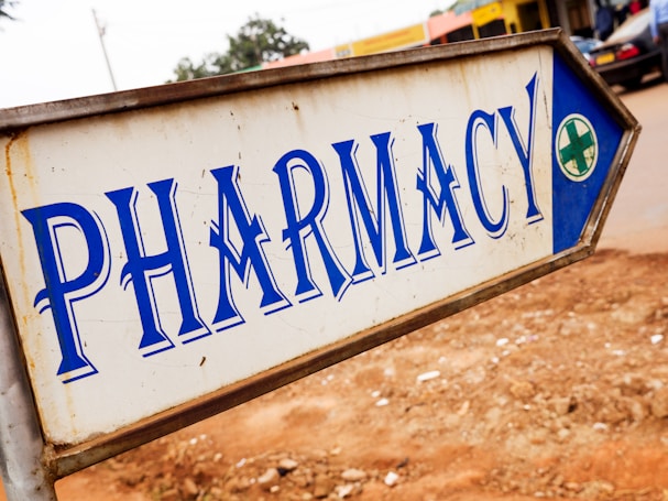 A metal sign with blue lettering indicating the direction to a pharmacy. The background is slightly blurred, showing pavement and some indistinct buildings and vehicles.
