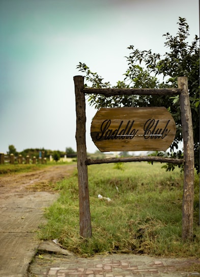 A rustic wooden sign marked 'Saddle Club' is positioned near a pathway surrounded by grass and greenery. The scene suggests an outdoor or rural setting, with a cloudy sky and wooden posts supporting the sign. In the background, there are additional wooden structures or fences leading into a field.