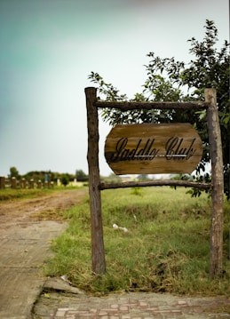 A rustic wooden sign marked 'Saddle Club' is positioned near a pathway surrounded by grass and greenery. The scene suggests an outdoor or rural setting, with a cloudy sky and wooden posts supporting the sign. In the background, there are additional wooden structures or fences leading into a field.