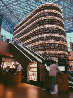 A large, multi-story bookshelf filled with books dominates the center of a spacious modern library. The structure is illuminated with warm lighting, creating a cozy yet grand atmosphere. An escalator on the left side allows people to access different levels of the bookshelf. Several people are engaged with their activities, including browsing and interacting with electronic kiosks. The ceiling features a geometric, patterned design with ample natural light streaming in through large windows.