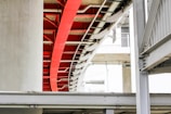 A modern architectural structure featuring intersecting metal beams and concrete pillars. The image highlights a prominent red beam amidst a predominantly white and grey color palette. The design includes curves and straight lines, creating an industrial yet artistic appeal.