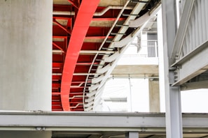 A modern architectural structure featuring intersecting metal beams and concrete pillars. The image highlights a prominent red beam amidst a predominantly white and grey color palette. The design includes curves and straight lines, creating an industrial yet artistic appeal.