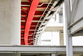 A modern architectural structure featuring intersecting metal beams and concrete pillars. The image highlights a prominent red beam amidst a predominantly white and grey color palette. The design includes curves and straight lines, creating an industrial yet artistic appeal.