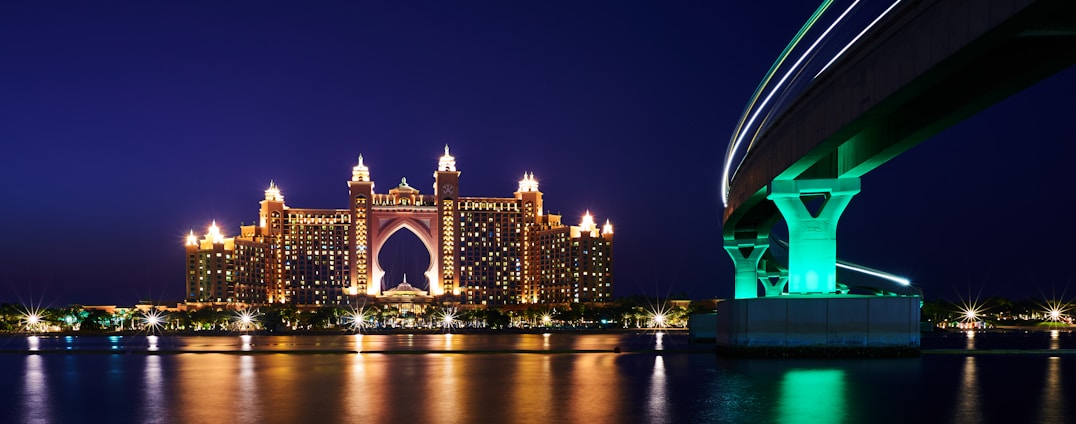 A grand, illuminated hotel with ornate towers reflecting in the calm water during nighttime. A modern bridge with a dramatic curve and glowing green lights is visible to the right, contributing to a striking architectural contrast.