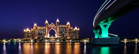 A grand, illuminated hotel with ornate towers reflecting in the calm water during nighttime. A modern bridge with a dramatic curve and glowing green lights is visible to the right, contributing to a striking architectural contrast.