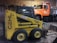 A yellow skid steer loader with the label 'GEHL 4625 SX' is parked inside a garage or workshop. There's an orange dump truck with the name 'Weston' visible in the background.