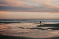 A sunset over a quiet beach with a lone traveler walking along the shore.