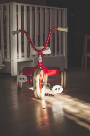 A red tricycle is placed on a wooden floor, illuminated by a soft beam of light. In the background, there is a white crib and a part of a chair or stool visible. The scene appears to be in a dimly lit room with a calm and nostalgic atmosphere.