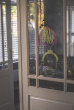 A child's drawing is visible on a glass panel door. The drawing appears to be made with yellow and red markers, featuring a simple face with scribbles around it. The background shows elements of a home interior with soft lighting streaming through partially opened blinds.
