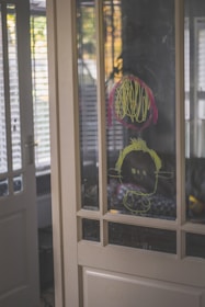 A child's drawing is visible on a glass panel door. The drawing appears to be made with yellow and red markers, featuring a simple face with scribbles around it. The background shows elements of a home interior with soft lighting streaming through partially opened blinds.