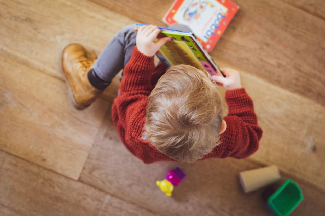 A young child with light blonde hair is wearing a red sweater and sitting on a wooden floor. The child is engaged in reading a colorful book. Nearby, there are a few colorful toys scattered around, including a yellow object, a purple object, and a green container.