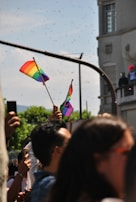 A crowd of people is gathered outdoors, some holding rainbow flags aloft. The setting conveys a sense of community and celebration, with a building and trees visible in the background.