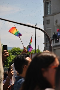 A crowd of people is gathered outdoors, some holding rainbow flags aloft. The setting conveys a sense of community and celebration, with a building and trees visible in the background.