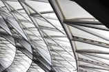 The image features the intricate architecture of a large, modern roof structure composed of steel beams and glass panels. The beams form a geometric pattern, creating an aesthetic interplay of lines and angles. Sunlight softly filters through the glass, creating a bright and airy ambiance.