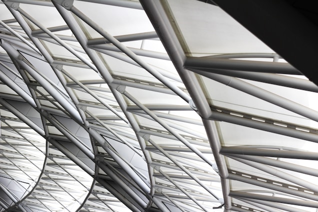 The image features the intricate architecture of a large, modern roof structure composed of steel beams and glass panels. The beams form a geometric pattern, creating an aesthetic interplay of lines and angles. Sunlight softly filters through the glass, creating a bright and airy ambiance.