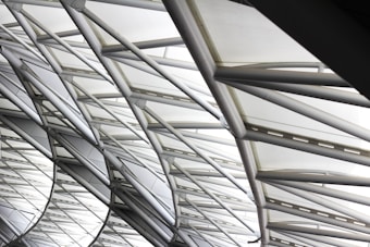 The image features the intricate architecture of a large, modern roof structure composed of steel beams and glass panels. The beams form a geometric pattern, creating an aesthetic interplay of lines and angles. Sunlight softly filters through the glass, creating a bright and airy ambiance.