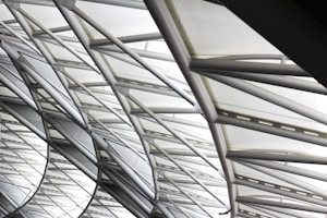The image features the intricate architecture of a large, modern roof structure composed of steel beams and glass panels. The beams form a geometric pattern, creating an aesthetic interplay of lines and angles. Sunlight softly filters through the glass, creating a bright and airy ambiance.