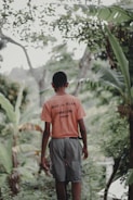 A person wearing a bright orange shirt and gray shorts walks along a lush, green path surrounded by tropical foliage. The back of their shirt features text, and they appear to be alone in a serene natural setting.