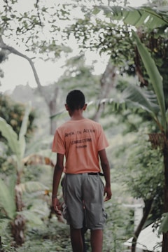 A person wearing a bright orange shirt and gray shorts walks along a lush, green path surrounded by tropical foliage. The back of their shirt features text, and they appear to be alone in a serene natural setting.
