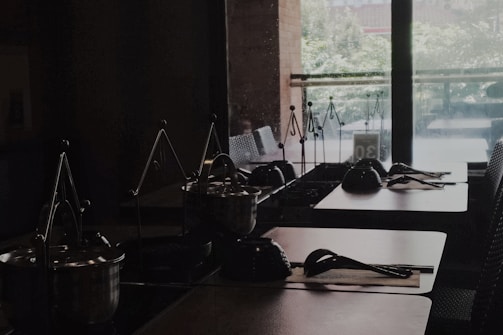 A dimly lit interior space with several tables arranged in a row, each set with pots on portable stoves. Metal trivets and serving utensils are neatly placed on the tables, and light filters in through large glass windows, offering a glimpse of greenery outside.