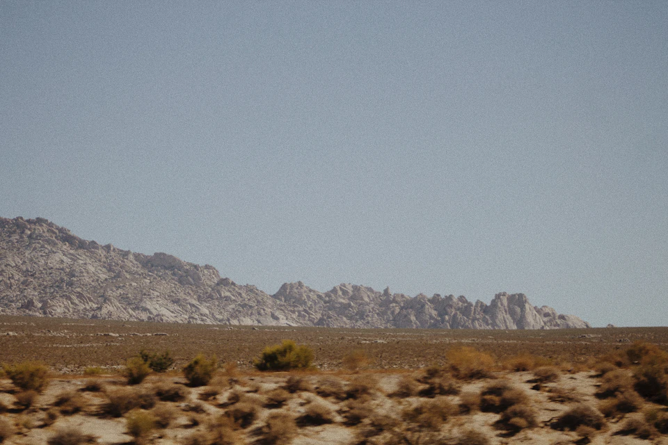 The image depicts a vast desert landscape with a clear blue sky and distant mountains. The mountains, covered in dry grass and shrubs, rise against the horizon, creating a sense of depth and scale. In the foreground, there are several small hills scattered across the scene, adding texture to the otherwise flat terrain. The predominant colors in the image are shades of brown from the desert vegetation and blue from the sky, with some green hues from the distant mountains.