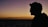 A silhouette of a person wearing glasses against a serene sunset backdrop with a calm ocean beneath a gradient sky of orange to purple.
