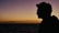 A silhouette of a person wearing glasses against a serene sunset backdrop with a calm ocean beneath a gradient sky of orange to purple.