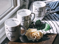Three white mugs with black script writing are placed on a wooden surface near a window. Each mug is inscribed with city names: Paris, Toronto, and a partially visible name that could be Los Angeles. Inside the rims are motivational phrases like 'Be Cool, Be Kind.' Accompanying the mugs is a single white rose on a striped cloth, with sunlight casting gentle shadows.