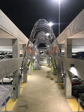 A modern multi-story parking garage at night, illuminated by artificial lighting. The structure features a prominent metallic sculpture with an intricate lattice design arching over a central walkway. Cars are parked on both sides, and signage for stairs is visible. Concrete pillars support the upper levels.
