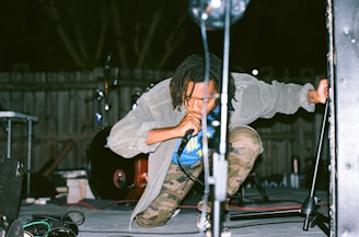 A person is crouched on stage holding a microphone, wearing a gray jacket and camo pants. The background features a fence and a drum set under dim lighting.