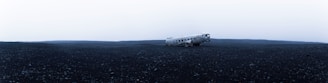 A desolate, wide-open landscape features a crashed airplane in the middle of a vast expanse of dark, rocky terrain. The sky above is overcast, creating a stark contrast between the dreary sky and the barren ground.