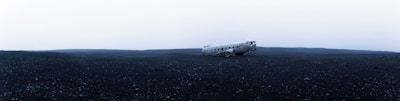 A desolate, wide-open landscape features a crashed airplane in the middle of a vast expanse of dark, rocky terrain. The sky above is overcast, creating a stark contrast between the dreary sky and the barren ground.
