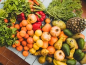 An assortment of fresh fruits and vegetables is arranged on a table. Among the produce are green leafy herbs, red bell peppers, oranges, lemons, pomegranates, pears, avocados, bananas, a pineapple, and more. The vibrant colors and natural textures create an inviting and healthy display.