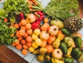 An assortment of fresh fruits and vegetables is arranged on a table. Among the produce are green leafy herbs, red bell peppers, oranges, lemons, pomegranates, pears, avocados, bananas, a pineapple, and more. The vibrant colors and natural textures create an inviting and healthy display.